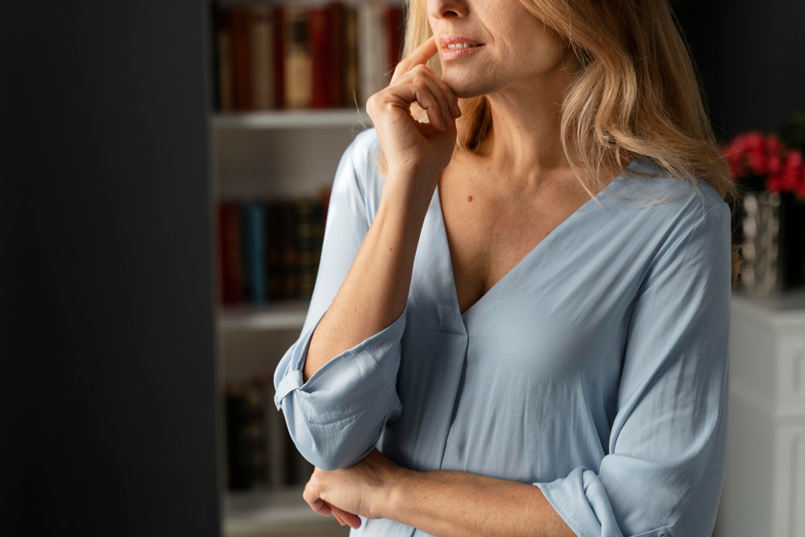 Middle-aged blonde woman in a blue blouse touching her chin thoughtfully with a bookshelf in the background.