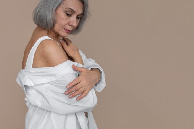 Older woman with short gray hair hugging herself with a white shirt slipping off her shoulder against a beige background.