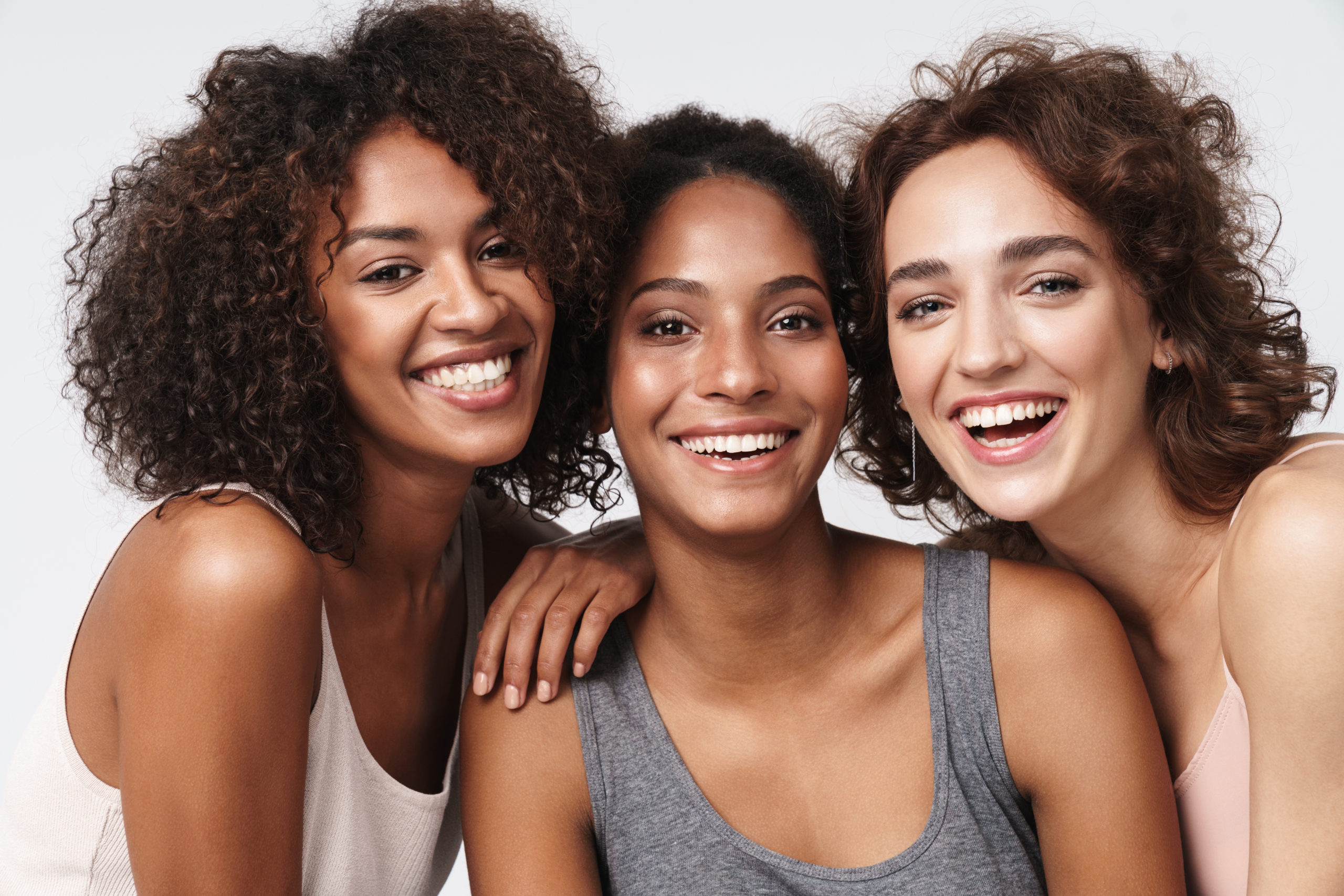 Portrait of three young multiracial women standing together and smiling at camera isolated over white background