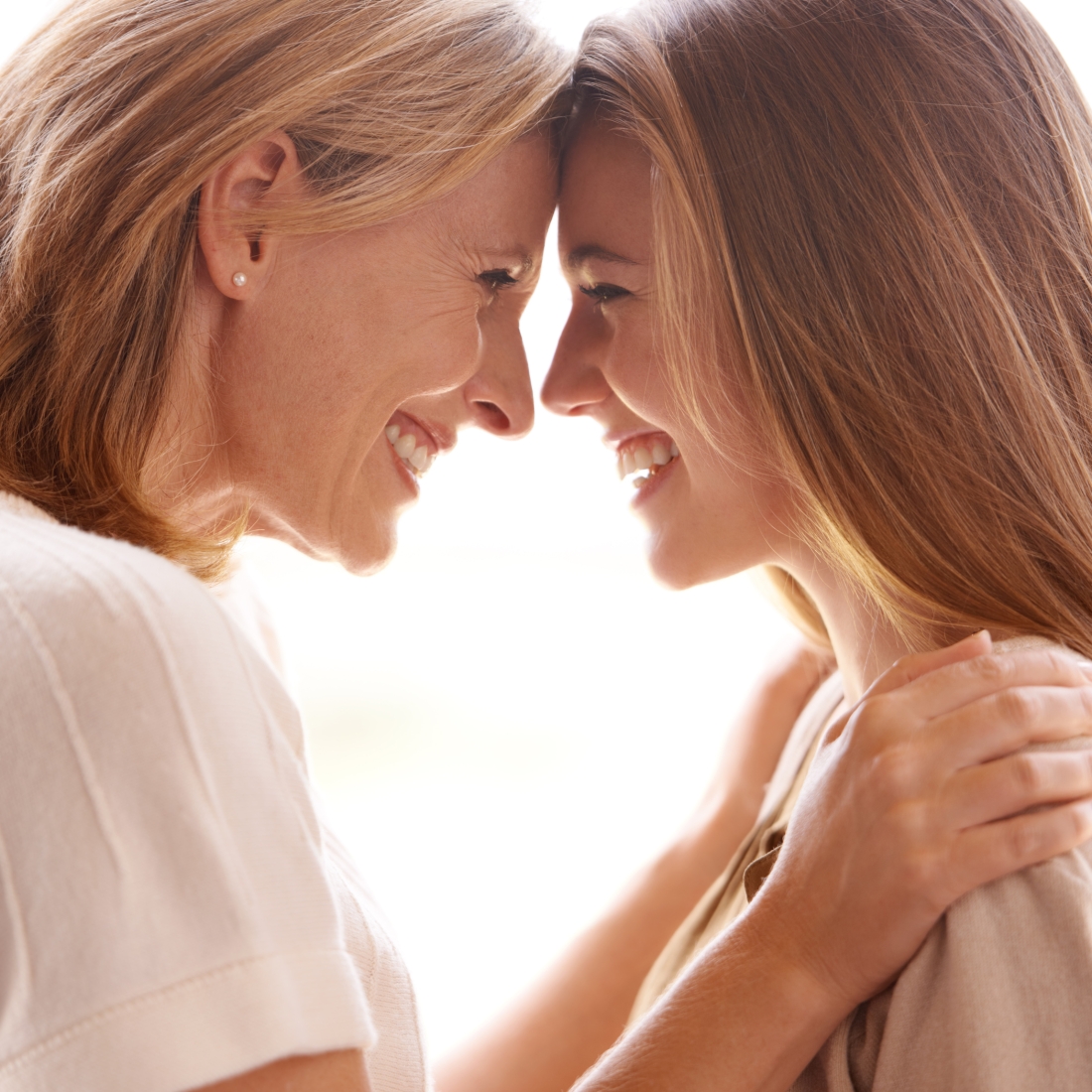 A close-up of a mother and daughter standing face to face, smiling warmly with their foreheads gently touching. Both women show deep affection and happiness, their hands resting softly on each other’s shoulders, captured in soft natural light that emphasises their loving bond.