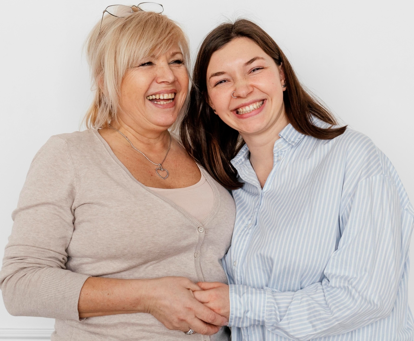 A middle-aged woman with blonde hair and a younger woman with long brown hair laughing heartily while holding hands. The mother wears a beige cardigan over a light top and a heart-shaped pendant necklace, and the daughter is dressed in a blue and white striped shirt with a nose ring. They stand against a plain white background, exuding joy and close family connection.