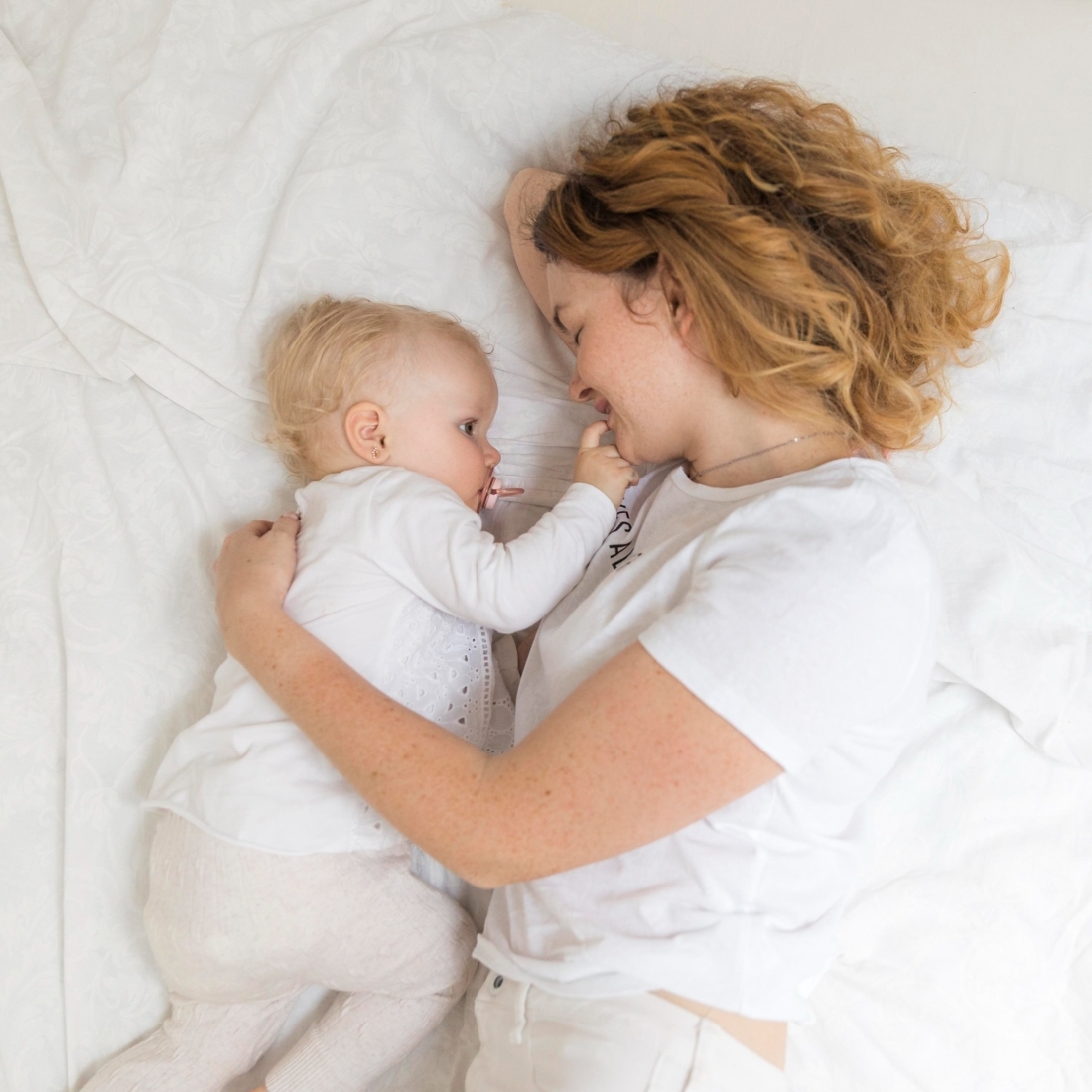 A tender moment of a mother with curly ginger hair lying on a bed, cuddling her blonde baby who has a pacifier. Both are dressed in white, sharing an affectionate and peaceful gaze.