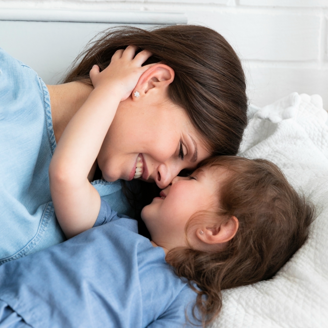 A close-up of a mother and daughter touching noses and smiling warmly at each other while lying down, sharing a loving and affectionate moment. Both are wearing blue tops, and the mother has dark brown hair, while the little girl has light brown curly hair.