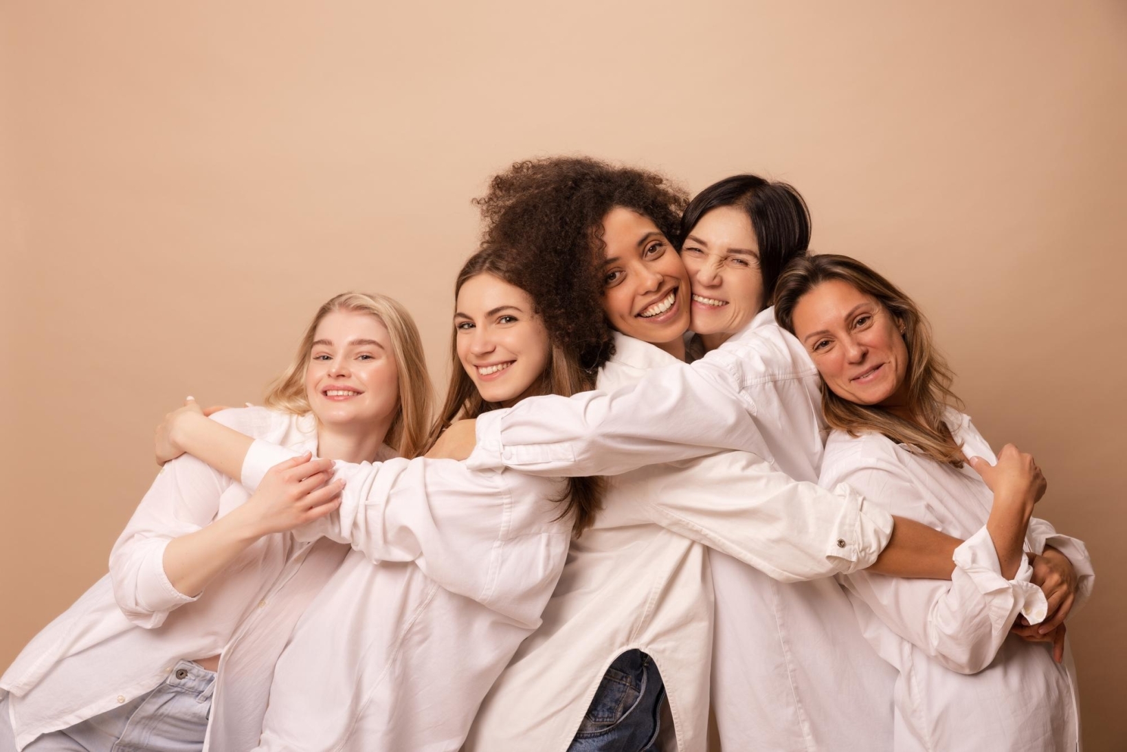 A group of five cheerful women standing close together, wearing white shirts and smiling warmly while hugging each other against a beige background.