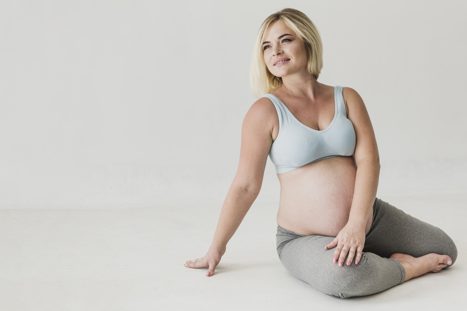 Pregnant woman with short blonde hair sitting on the floor in a relaxed pose, wearing a light blue sports bra and grey leggings, looking content.