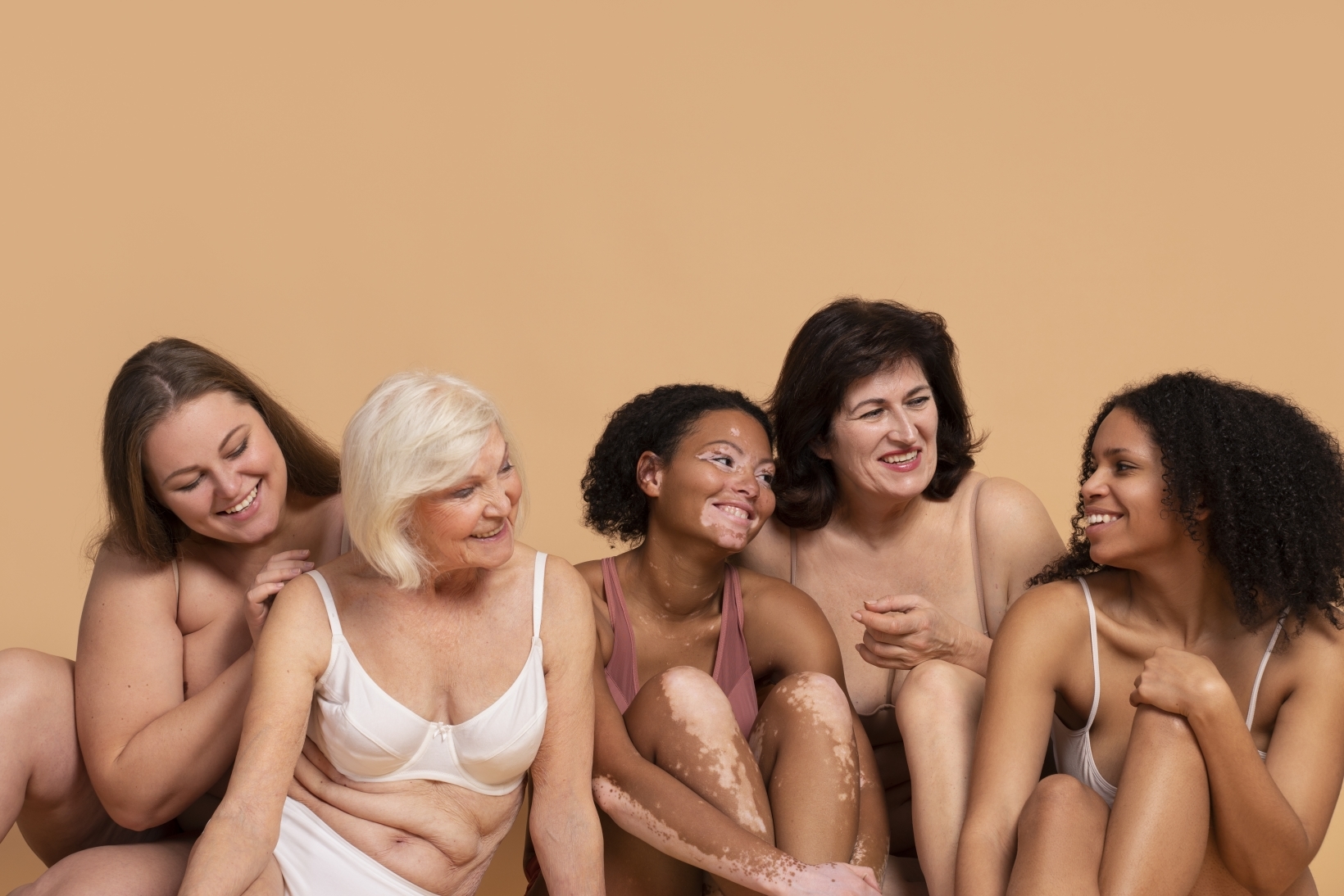 Group of diverse women of different ages and body types sitting together in underwear, smiling and enjoying each other's company against a beige background.