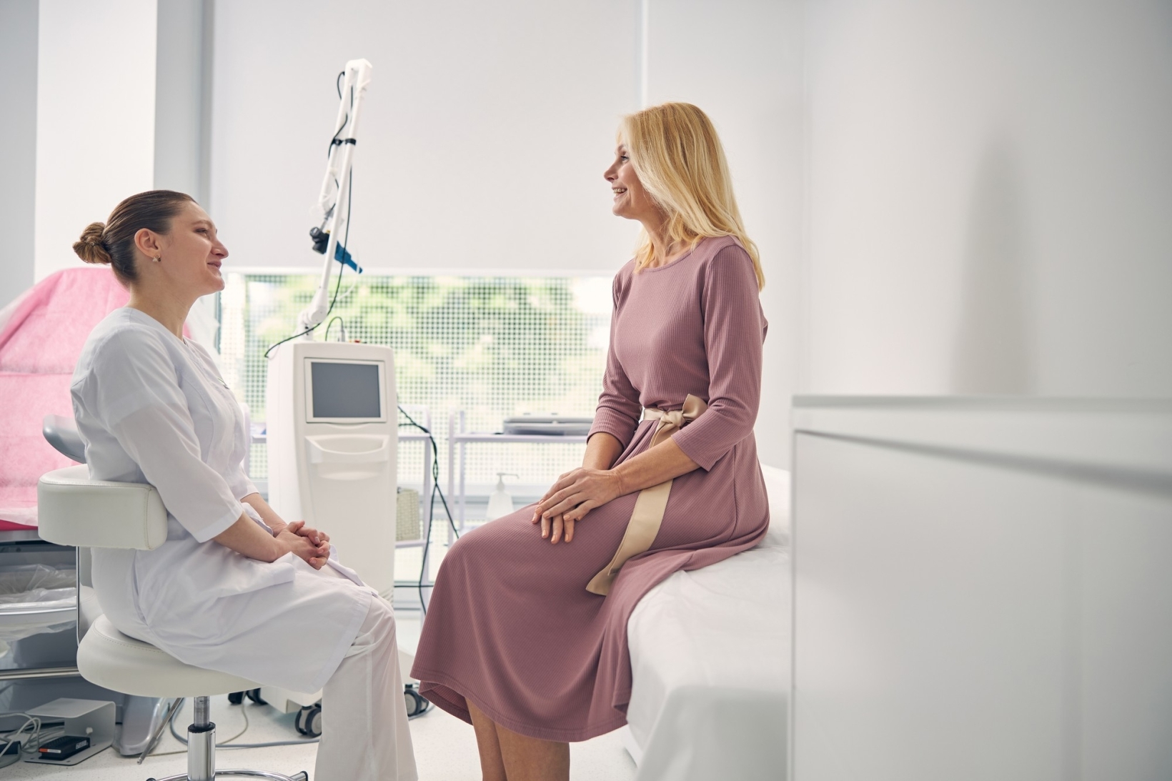 A female doctor in a white medical uniform sits on a stool while speaking with a woman in a long pink dress who is seated on an examination bed inside a bright, modern clinic.