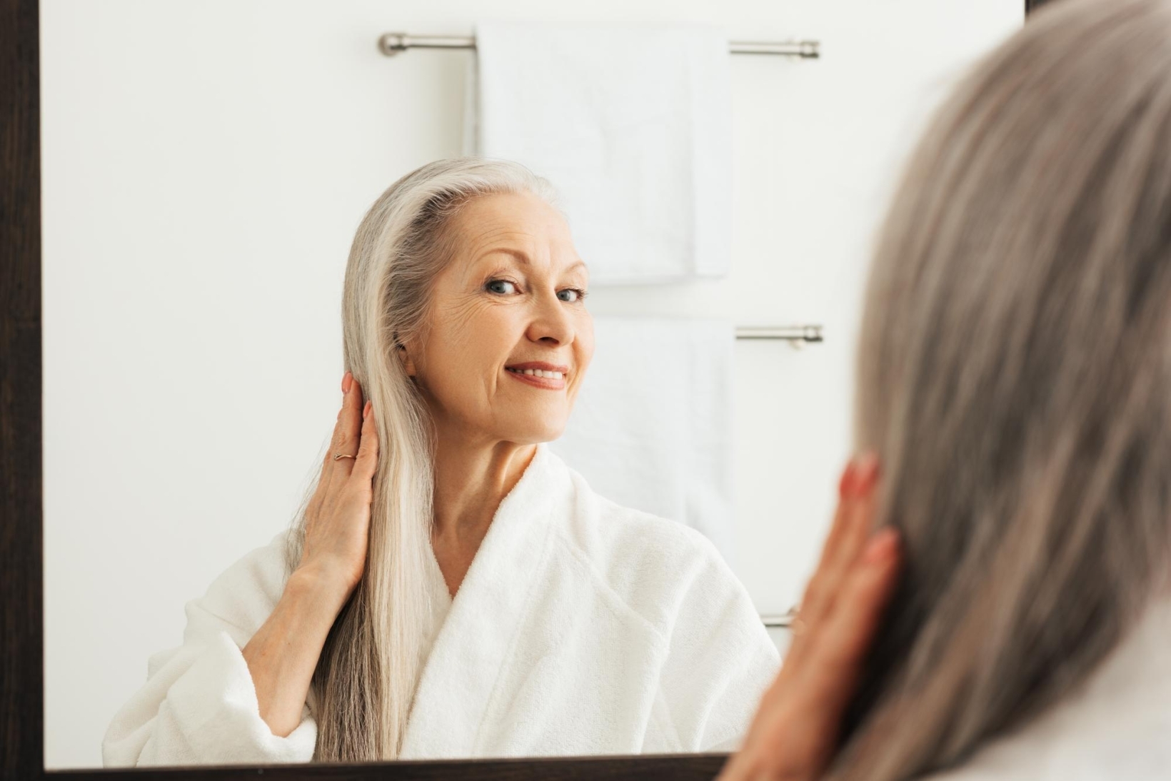 An older woman with grey hair smiling and adjusting her hair in a mirror in a bathroom.