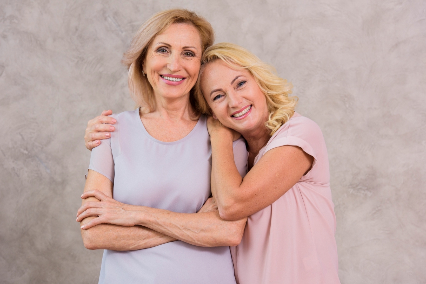 Two smiling older women standing close, one with her arm around the other, posing affectionately against a neutral background.