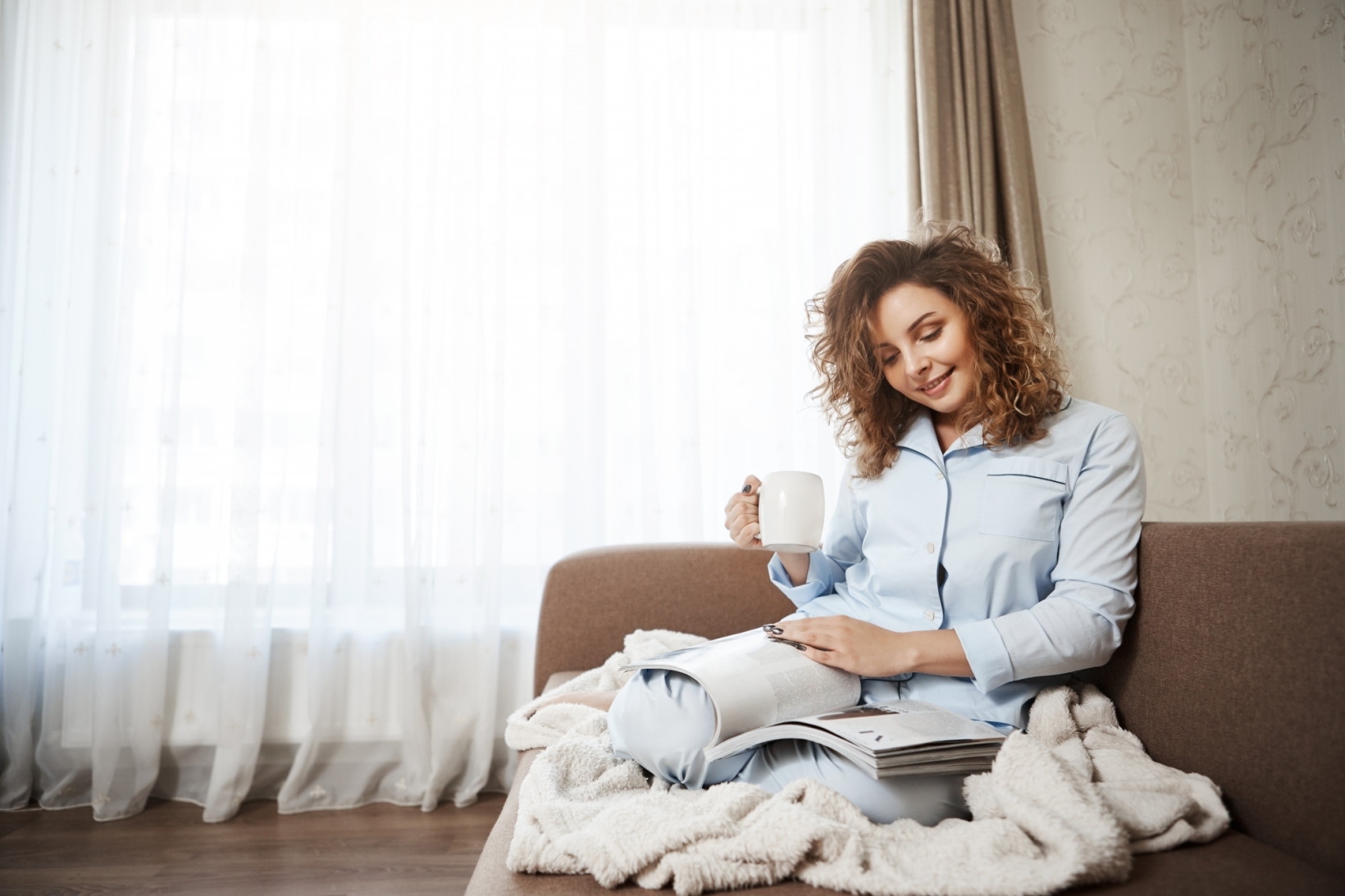 A woman in light blue pajamas is sitting on a sofa with a blanket, holding a coffee mug and reading a magazine near a bright window.