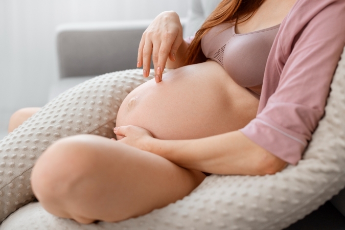 Close-up of a pregnant belly as a woman relaxes against supportive pillows, lightly touching her baby bump in a serene home setting.