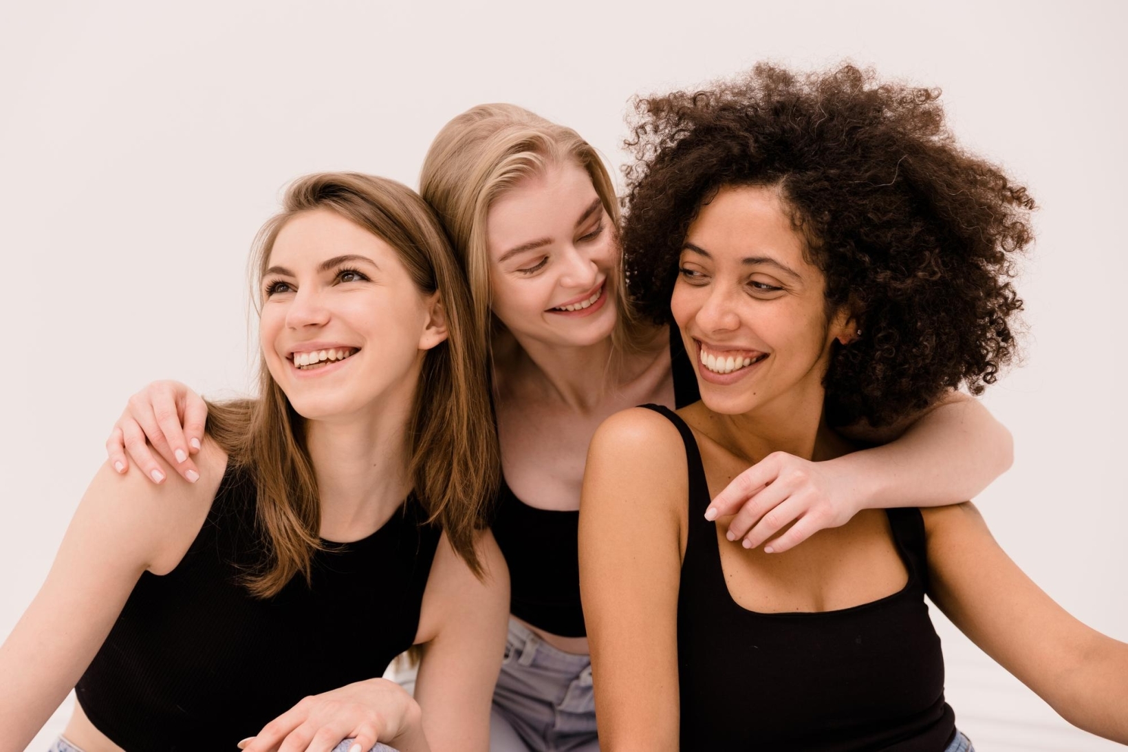 Three women of different backgrounds smiling and leaning close together, sharing a joyful moment in a bright studio setting.