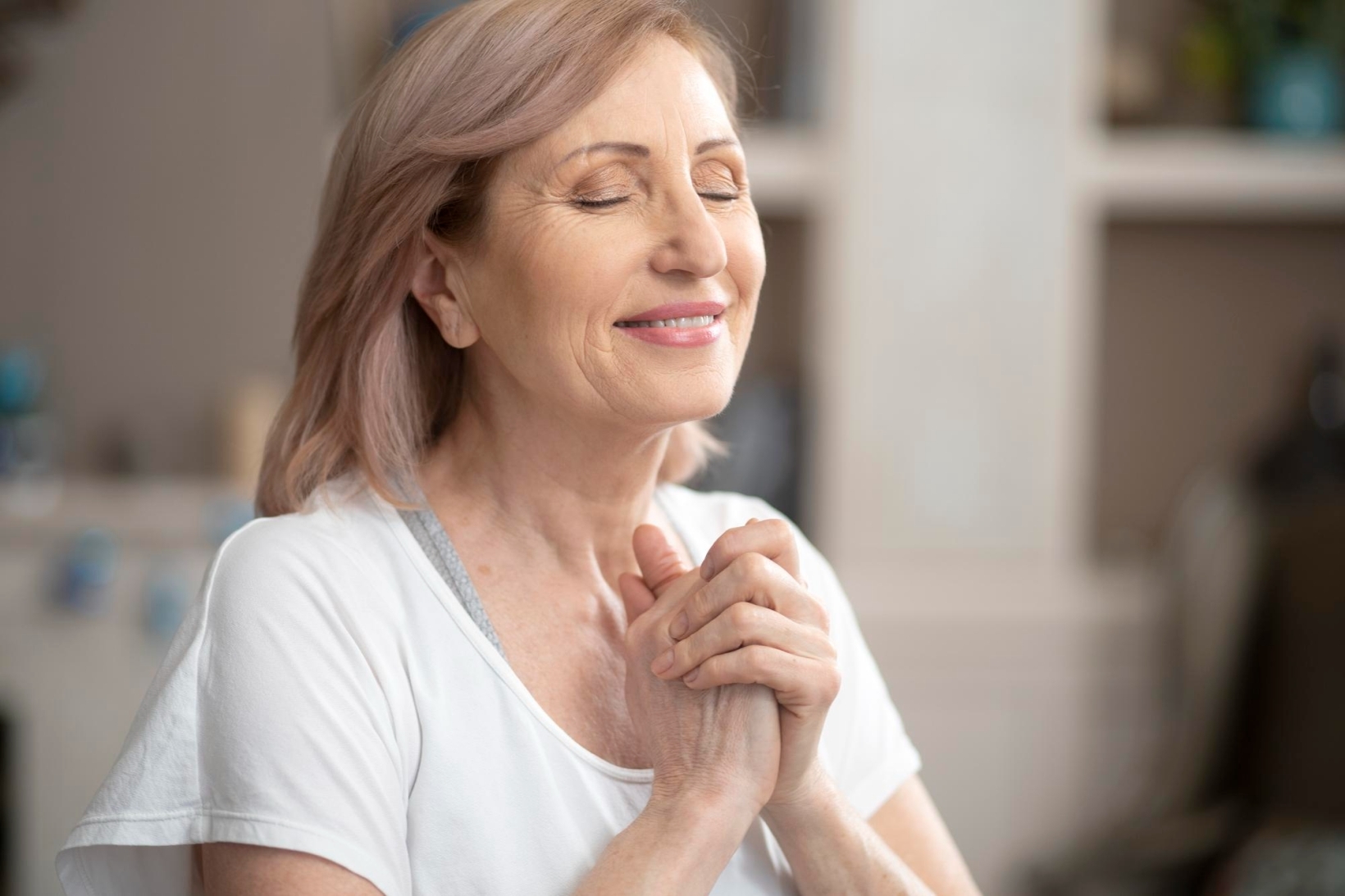 Smiling older woman with eyes closed and hands clasped together, appearing calm and content in a cozy home environment.