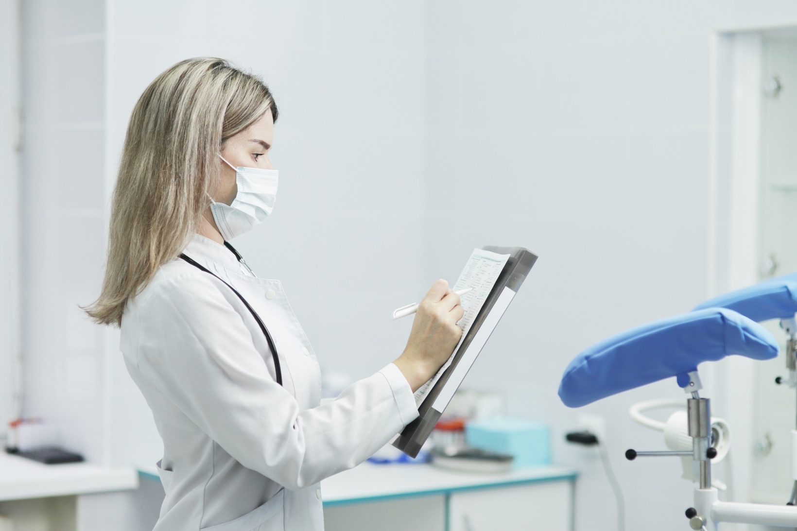Female gynecologist in a white coat and mask writing on a clipboard in an exam room.