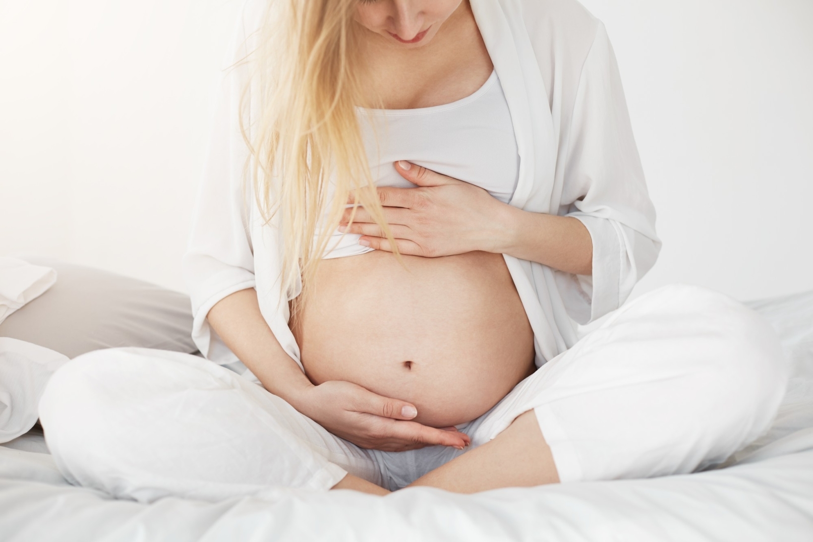 Pregnant woman sitting cross-legged on a bed holding her bare belly.