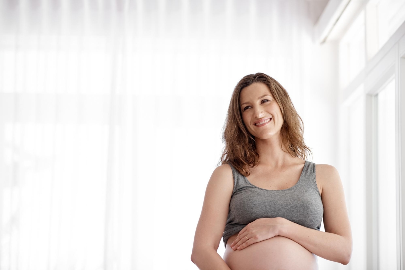 Smiling pregnant woman in a gray tank top holding her belly by a bright window.