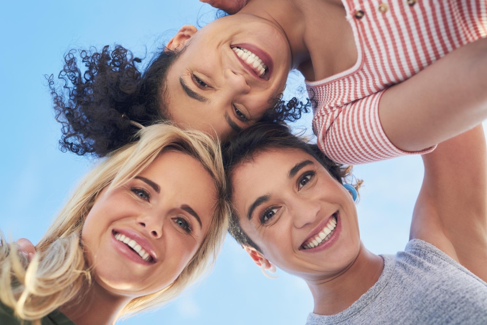Low-angle view of three smiling women huddled together against a blue sky.