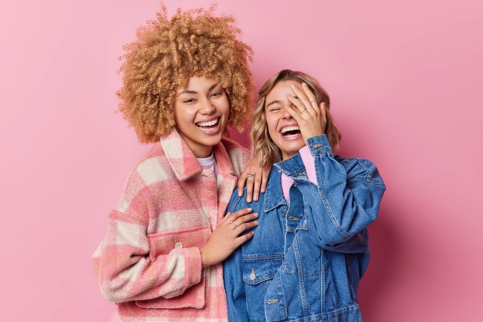 Two women laughing together against a pink background.