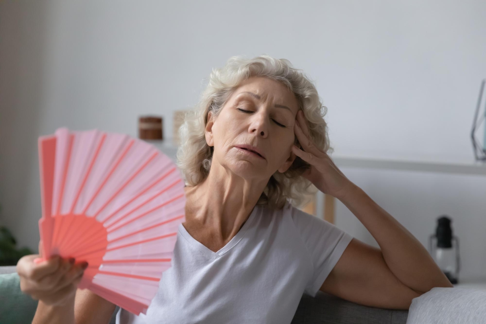 Older woman with white hair fanning herself with a pink hand fan while experiencing a hot flash.