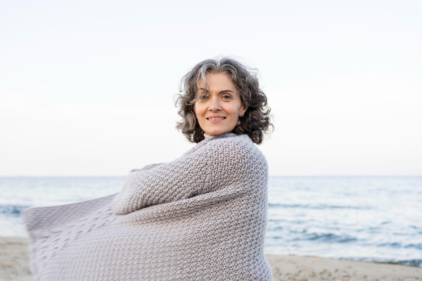 Smiling mature woman with curly grey hair wrapped in a knitted blanket by the sea