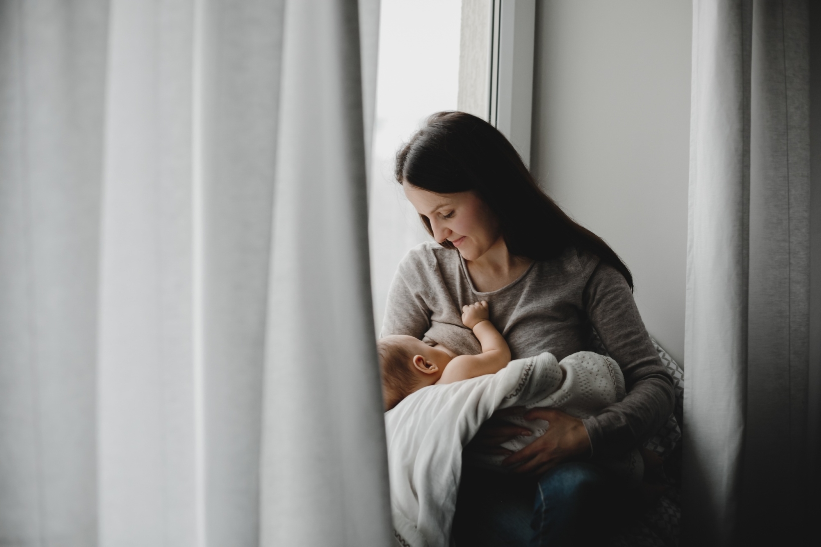 Mother sitting by a window cradling and breastfeeding her baby in soft natural light