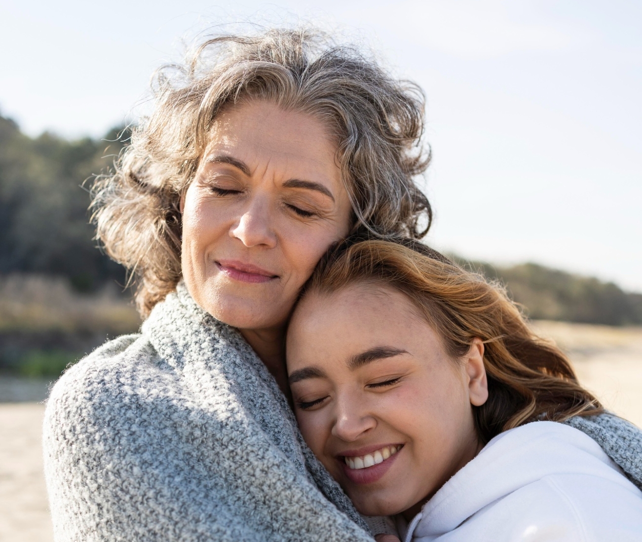 Older woman and younger woman wrapped together in a grey blanket, embracing with their eyes closed on a sunny beach