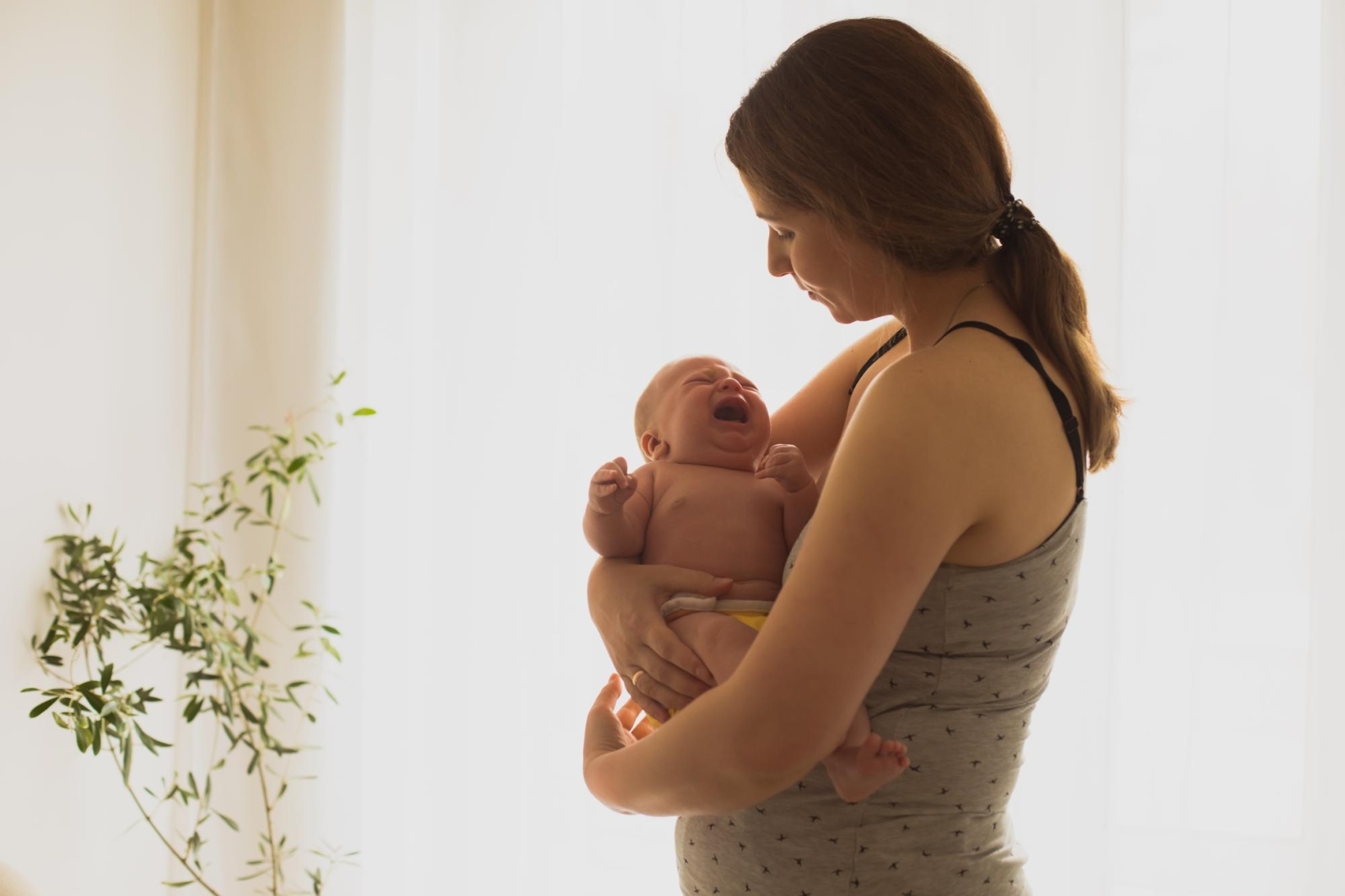 Mother standing by a bright window holding her crying newborn baby