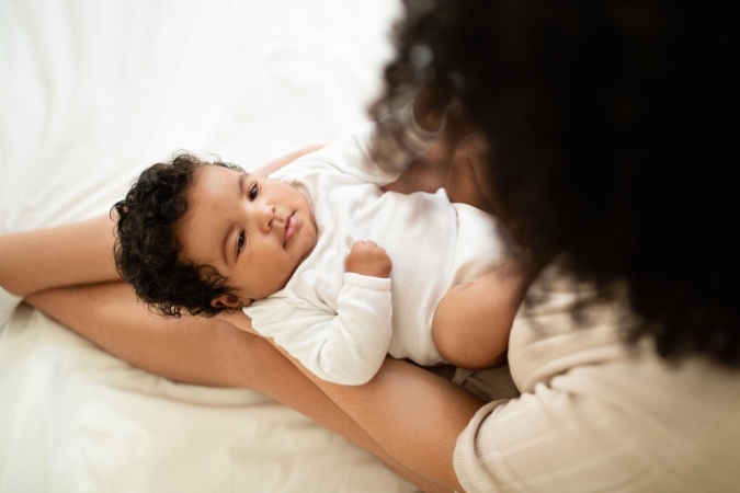 Overhead view of a curly-haired baby in a white bodysuit lying in its mother's arms on a bed, looking up at her