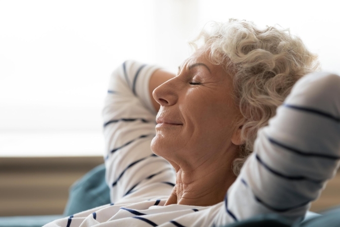 Senior woman with curly grey hair leaning back with her hands behind her head and eyes closed, relaxing in bright light