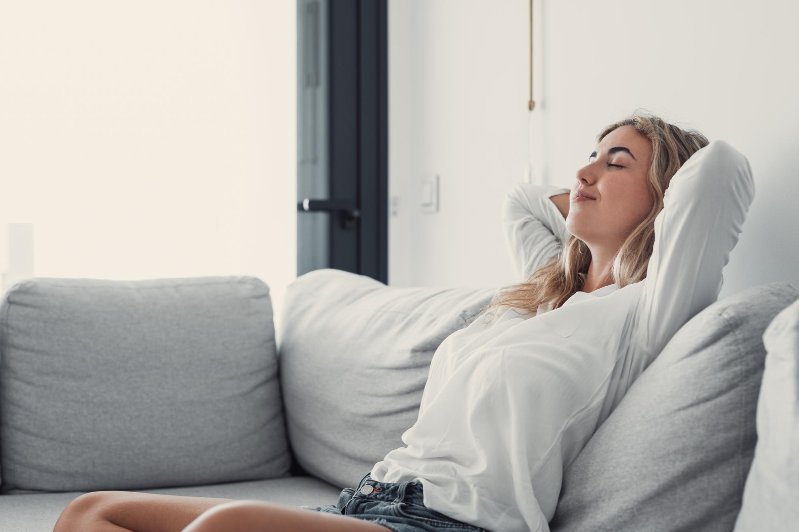 Young woman in a white shirt resting on a grey sofa with her hands behind her head and eyes closed